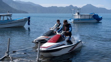 Two riders slowly guiding personal watercraft toward a trailer at a calm lakeside launch, demonstrating real-world jet ski loading conditions and alignment challenges